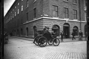 Två män åker i en äldre bil år 1900 medan människor står utmed gatan och kollar på.